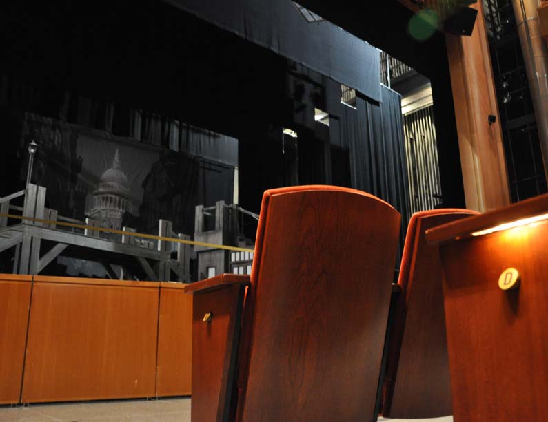 Empty theater seats facing a stage with dark curtains and a cityscape backdrop.